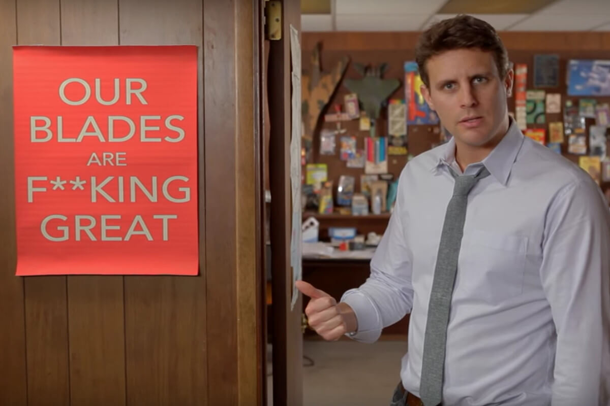 screenshot from dollar shave club ad with guy pointing to poster that reads "our blades are f**king great"